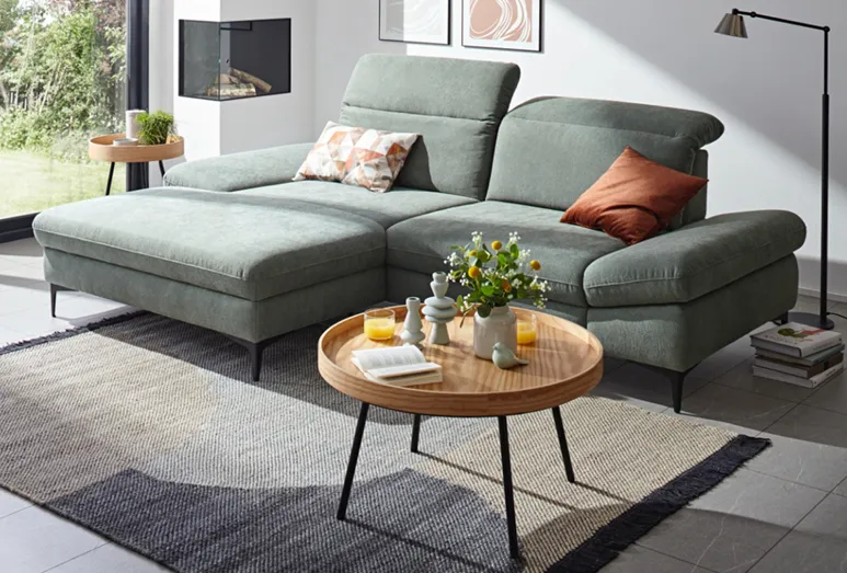 Global Alegria, ein ergonomisches Sofa in Grün mit verstellbarer, hoher Rückenlehne, dazu ein dekorativer runder Couchtisch in Holzoptik im hellen Wohnzimmer