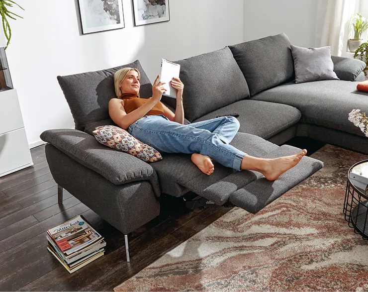 Blonde Frau liegt auf einem modernen dunkelgrauen Sofa und liest ein Buch.