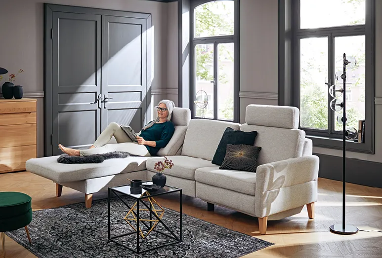 Frau lesend auf heller Global Arima Sofa im Wohnzimmer, im Hintergrund eine graue Tür im Landhausstil, vor dem Sofa ein Teppich auf dem Holzboden