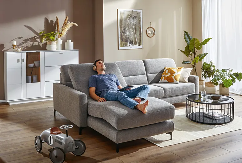 Frau mit Tasse Kaffee und Buch auf einer grauen Couch mit verstellbarer Rückenlehne, Mann mit Kopfhörern auf einer grauen Couch mit verstellbaren, ergonomischen Komponenten