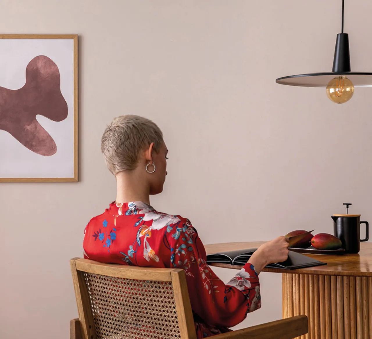 Frau sitzt an einem Holztisch und liest in einem Buch. Moderne Einrichtung mit hellen Wänden, minimalistischer Beleuchtung und stilvollem Kunstbild. Ruhige Wohnatmosphäre mit warmem, natürlichem Charakter.