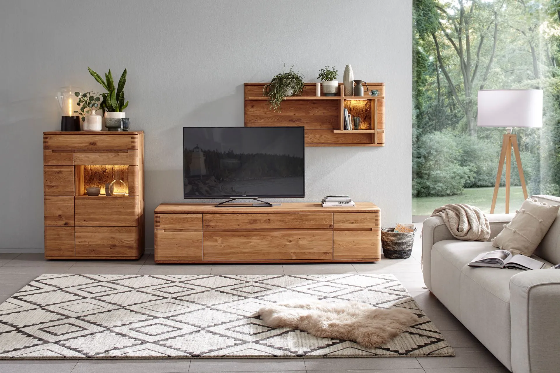 Helle Wohnwand aus Holz mit Glaselement im Highboard vor einer Wand neben einem Fenster.
