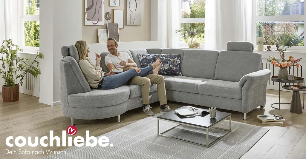 Modernes graues Ecksofa von Couchliebe in hell eingerichtetem Wohnzimmer, auf dem ein Paar entspannt sitzt – erhältlich bei Möbel Rommel.