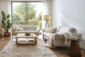 Zwei beige Sofas mit hellen Kissen und Decke in einem hellen Wohnzimmer mit großer Pflanze, Stehlampe und Couchtisch.