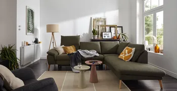 Grünes Ecksofa mit klappbaren Kopflehnen und hellen Holzfüßen, dekoriert mit mehreren Kissen und Decken in einem hellen gemütlichen Raum.