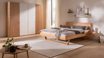 Helles gemütliches Schlafzimmer mit Kleiderschrank in hellem Holz mit Lamellenoptik und weißen Elementen an den Türen sowie ein dazu passendes Bett mit Lamellenoptik und weißen Elementen.
