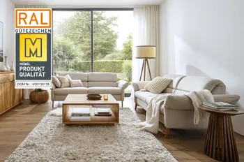 Gütesiegel &quot;Goldene M&quot; auf einem Bild von zwei beigen Sofas mit hellen Kissen und Decke in einem hellen Wohnzimmer mit großer Pflanze, Stehlampe und Couchtisch.