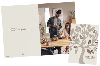 Aufgeschlagene Seite des neuen Natura Home Katalogs mit dem Schriftzug &quot;The home of harmony&quot; auf beigem Hintergrund, daneben ein Bild eines entspannten Paares am Esstisch und rechts daneben das Cover des Katalogs in brauntönen mit einem Baum.