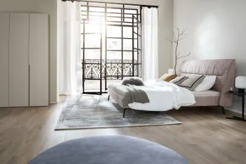 Seitliche Aufnahme eines modernen beigen Polsterbett, auf dem mehrere Kissen in beige, weiß, gelb und grau liegen sowie zwei Decken in weiß und grün.