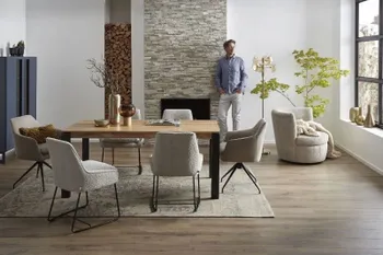 Modernes Esszimmer mit einem großen Esstisch aus Massivholz, um den sechs graue Stühle mit schwarzem Metallgestell stehen, im Hintergrund lehnt ein Mann an einer Wand.