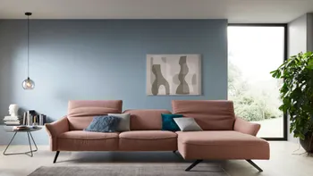 Rosesfarbenes Ecksofa mit modernen schwarzen Füßen in einem hellen Wohnzimmer mit blauer Wand im Hintergrund neben einem großen Fenster.
