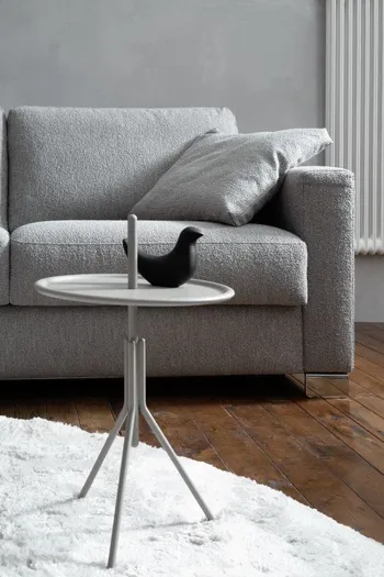 Rechte Ecke eines grauen Sofas mit großen grauen Rückenkissen und einem grauen Dekokissen, davor steht ein runder kleiner Bestelltisch mit einem schwarzen Dekovogel.