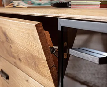 Detailaufnahme einer geöffneten Holztür mit moderner Klappfunktion an einem Sideboard aus massiver Eiche – eleganter Materialmix aus Holz und Glas mit schwarzen Metallgriffen.