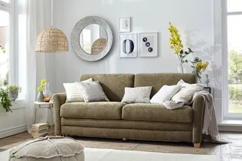 Grünes Sofa mit Schlafoption, auf dem weiße und beige Kissen und eine graue Decke liegen, über dem Sofa hängen Bilder und eine Spiegel an der Wand.