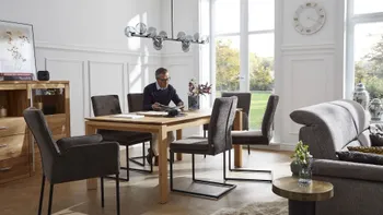 Mehrere graue Freischwinger mit schwarzem Metallgestell und zwei graue Armlehnstühle um einen Holz Esstisch, auf einem der Stühle sitzt ein Mann und liest.