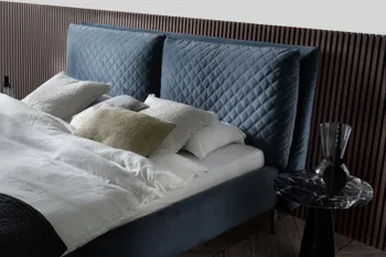 Kopfteil eines Boxspringbettes mit blauem Bezug, das Kopfteil besteht aus zwei Polstern mit Rautenmustern, auf dem Bett liegen Kissen und Decken in beige und weiß.