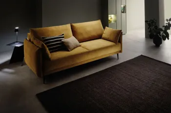 Cognac farbenes modernes Zweisitzer Sofa in einem dunklen Raum, neben dem Sofa steht ein schwarzer Beistelltisch mit einer kleinen Tischlampe, die angeschalten ist.