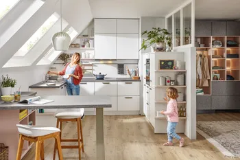 Mutter mit Tochter in kompakter Winkelküche in Dachgeschosswohnung mit weißen Küchenfronten. 