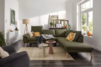 Grünes Ecksofa mit klappbaren Kopflehnen und hellen Holzfüßen, dekoriert mit mehreren Kissen und Decken in einem hellen gemütlichen Raum.