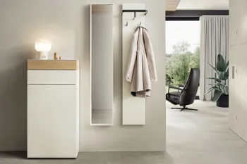 Weiße moderne Design Garderobenmöbel bestehend aus Highboard mit Holzelement, schmalem hängenden Spiegelschrank und hängendem weißen Brett mit schwarzen Garderobenhaken an dem ein Trenchcoat hängt.