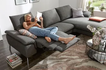 Eine Frau liegt gemütlich auf einem grauen Ecksofa mit ausgeklappter Fußstütze.