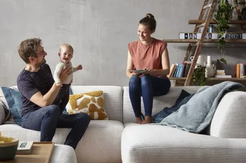 Mann mit einem Baby auf dem Schoss sitzt auf einem beigen Cordsofa, daneben sitzt eine lächelnde Frau auf der Rückenlehne des Sofas.
