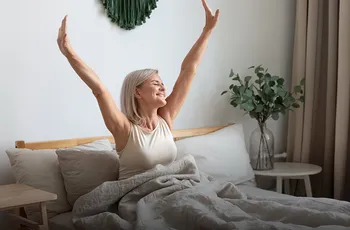 Frau sitzt auf einem Bett und reckt entspannt und zufrieden die Arme in die Luft.