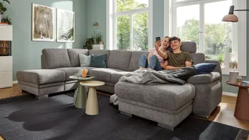 Graue Wohnlandschaft mit silbernen Füßen, auf dem ein Junge und ein Mädchen nebeneinander entspannt auf dem Longchair sitzen.