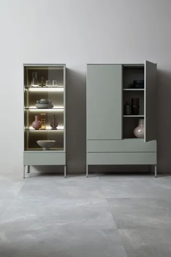 Salbeigrünes Highboard neben einer Salbeigrünen Vitrine mit beleuchteten Regalböden.