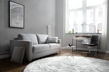 Hellgraues Zweisitzer Sofa mit grauem Kissen in einem hellen Raum mit großen Fenstern, vor denen  Vorhänge hängen.