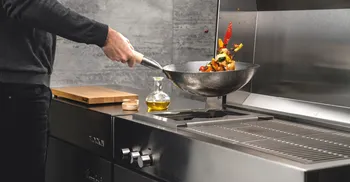 Mann schwingt Wokpfanne mit verschiedenem Gemuese ueber Heizflaeche einer Outdoorkueche. 