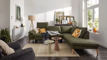 Grünes Ecksofa mit klappbaren Kopflehnen und hellen Holzfüßen, dekoriert mit mehreren Kissen und Decken in einem hellen gemütlichen Raum.