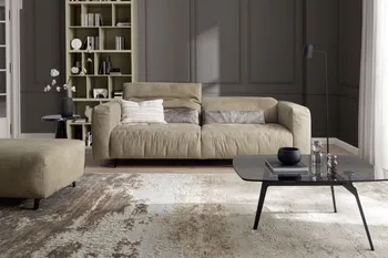 Modernes Ledersofa in Taupe mit aufgestellter Kopfstütze in einem modernen Wohnzimmer mit passendem Hocker und Glas Couchtisch in schwarz.