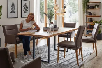 Helles Esszimmer mit Esstisch aus Massivholz mit Baumkante, braunen Lederstühlen und Armlehnstühlen und brauner Sitzbank, auf der eine Frau gemütlich sitzt.