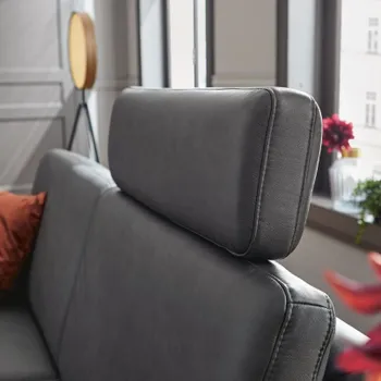 Graues Ledersofa mit Kopfstütze und rotem Kissen auf der Sitzfläche vor großen Fenstern.