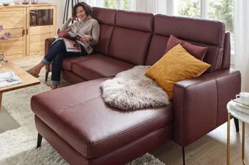 Weinrotes Ecksofa mit hoher Rückenlehne, auf dem eine ältere Frau sitzt und eine Zeitschrift anschaut.