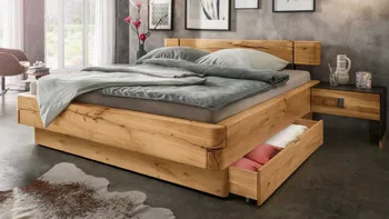 Massivholzbett mit abgerundeten Ecken das mit grauem und grünem Bettzeug dekoriert ist und einer geöffneten Schublade unterm Bett, mit passendem am Bett befestigten Couchtisch, der einen schwarzen Rahmen hat.