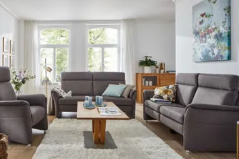 Zwei graue Sofas mit hohen Rückenlehnen, die im rechten Winkel zueinander stehen, gegenüber noch ein passender grauer Sessel in einem hellen Wohnzimmer.
