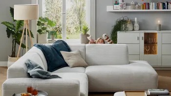 Hellgraues Ecksofa aus Cord, dekoriert mit mehreren Kissen, hinter der Rückenlehne sieht man ein Kind, das mit Handpuppen spielt.