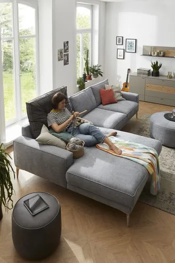 Hellblaues Ecksofa mit einem großen schwarzen Rückenkissen beim Longchair, auf dem eine Frau entspannt liegt und strickt.