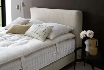 Boxspringbett mit beigem Boucle Bezug auf der Matratze liegt ein Topper und darauf mehrere Kissen in weiß und beige.