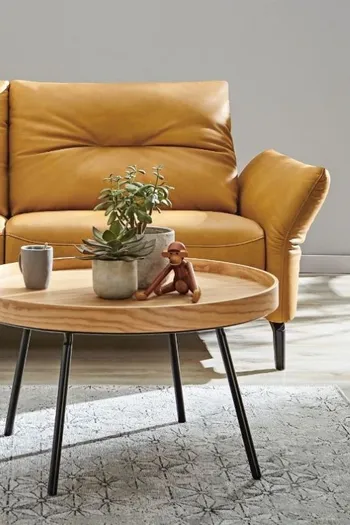 Rechter Teil eines gelben Ledersofas mit dick gepolsterten Rückenlehnen, davor steht ein runter Beistelltisch mit Holztischplatte.
