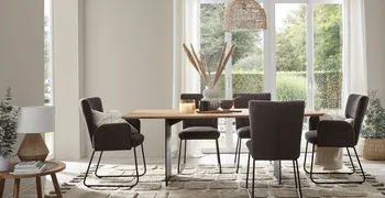Graue Polsterstühle mit schwarzem Metallgestell an einem Esstisch aus Holz, der dekoriert ist mit Vasen, eine mit großen Trockenblumen darin und Tassen und Schalen.