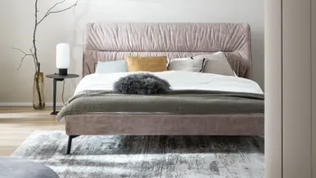 Beiges Design Polsterbett mit einer Querfalte am Kopfteil und mehreren Kissen in beige, gelb, und schwarz und Decken in weiß und grün.