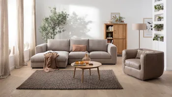 Graues Ecksofa mit großen grauen Kissen als Rückenpolster, dekoriert mit einer Decke und Kissen in rose, daneben ein passender Sessel in braunem Leder.