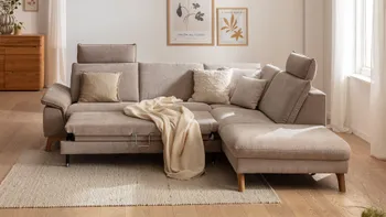 Beiges Ecksofa mit Holzbeinen, bei dem die Sitzfläche ausgezogen ist, auf dem Sofa liegen beige Kissen und eine beige Decke.