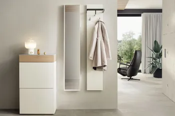 Weiße moderne Design Garderobenmöbel bestehend aus Highboard mit Holzelement, schmalem hängenden Spiegelschrank und hängendem weißen Brett mit schwarzen Garderobenhaken an dem ein Trenchcoat hängt.
