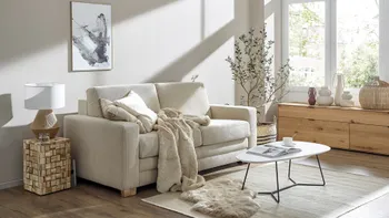 Beiges Zweisitzer Sofa in einem hellen Wohnzimmer, das mit einer Decke und Kissen in beige gemütlich dekoriert ist.