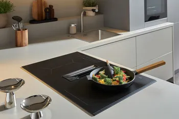 Kochfeld einer grauen Designküche, auf der eine Pfanne mit Gemüse steht.