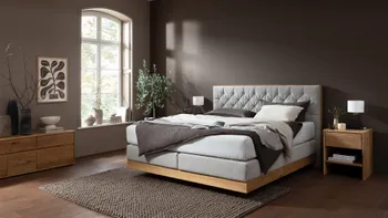 Graues Boxspringbett mit gestepptem Kopfteil mit hellem und dunklen Kissen und Decken dekoriert vor einer braunen Wand in einem gemütlichen Schlafzimmer.